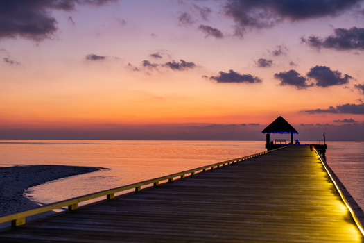 Wooden pier with yellow railings extending into calm water at dusk, illuminated boathouse at the end, purple and orange sunset sky with clouds.