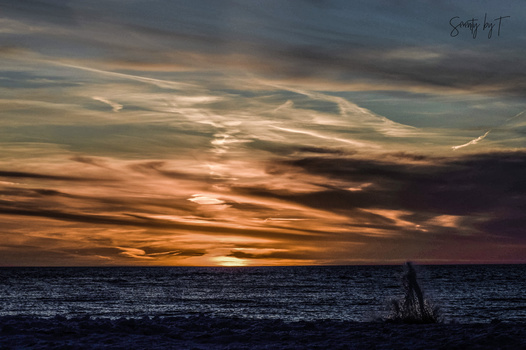 Silhouetted figure wading in ocean at dramatic sunset with stratified clouds.