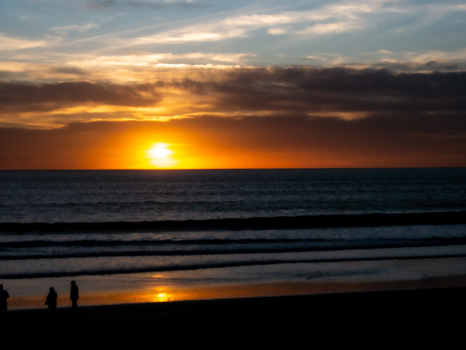 Golden sun setting over the ocean horizon with dramatic orange and dark clouds reflected in wet sand below.