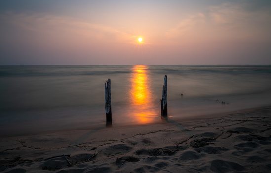 Sun reflecting on calm ocean water at sunset, with two weathered wooden posts in the shallow surf.