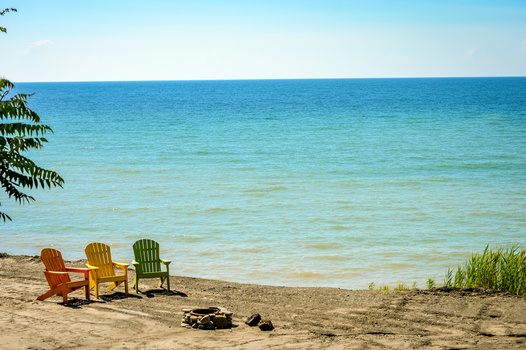 Three colorful Adirondack chairs on a sandy beach facing calm turquoise water.