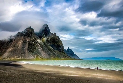 Dramatic volcanic mountain peaks rising behind a black sand beach and turquoise lagoon under stormy skies.