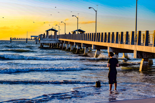 Person wading in shallow ocean water at sunrise with a long pier extending into the sea.