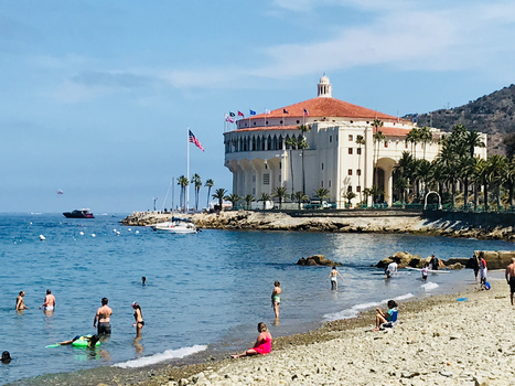 Beachgoers swim and wade in turquoise water near a historic waterfront castle with red-tile roof.