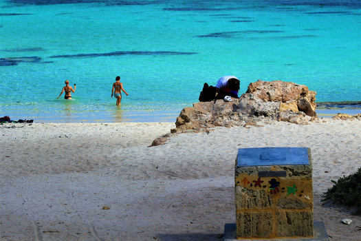 Turquoise Mediterranean beach with swimmers, rocky outcrops, and a weathered painted box in the foreground.