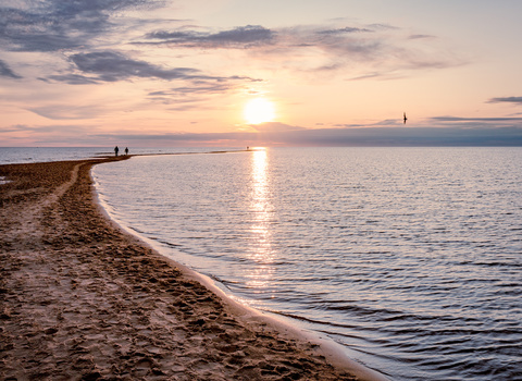 Sandy beach curving along calm water at sunset, with figures visible on the shore and sailboat on the horizon.