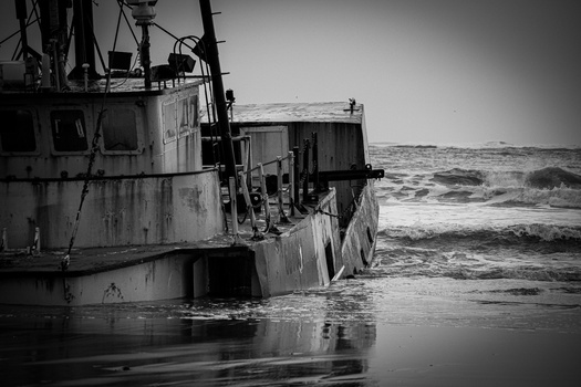 Abandoned tugboat tilted in shallow ocean waves, rendered in black and white.