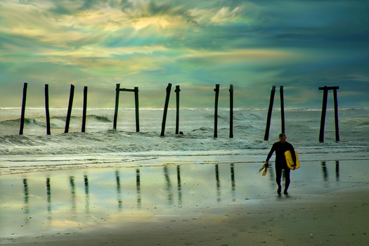 Surfer carrying a yellow and black board walking toward the ocean with weathered wooden pier pilings in the background.