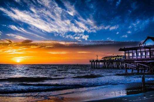 Wooden pier extending into the ocean during golden hour sunset with dramatic orange and blue sky.