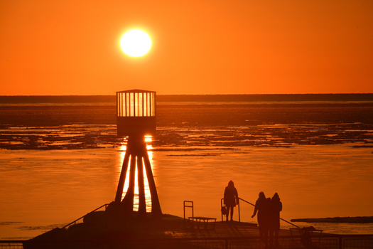 Silhouetted lighthouse and figures on a pier at sunset with golden sky and reflective water.