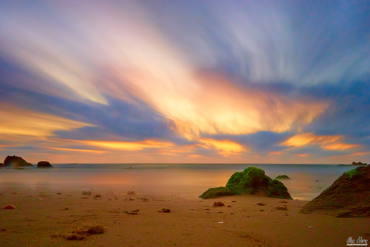 Beach at sunset with golden sky, scattered rocks, and calm ocean under dramatic clouds.