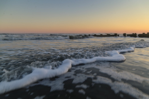 Ocean waves breaking on a dark sand beach at sunset with buildings visible along the shoreline.