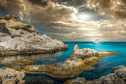 Rocky limestone formations in turquoise Mediterranean waters with a sailing yacht anchored in the distance.