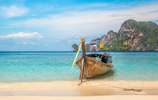 Wooden longtail boat with yellow and blue fabric moored in turquoise shallow water near a sandy beach with limestone cliffs.