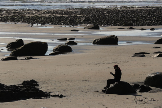 Silhouetted figure sitting on a rock amid scattered boulders on a wide beach at low tide.