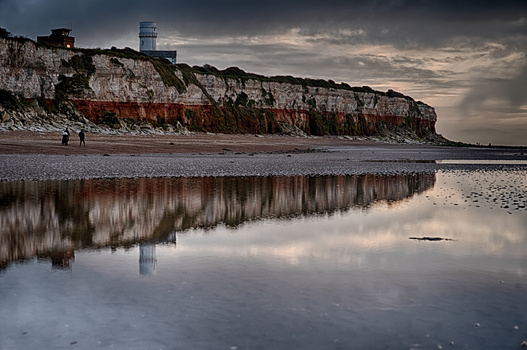 Dramatic coastal cliff with red strata reflected in still water at low tide.