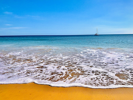 Golden sand beach with turquoise water and white waves, sailboat visible on the horizon under clear blue sky.