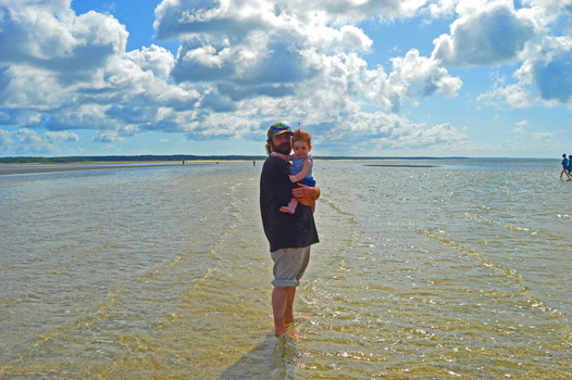 Man wading in shallow ocean water while holding a young child in an orange life vest.