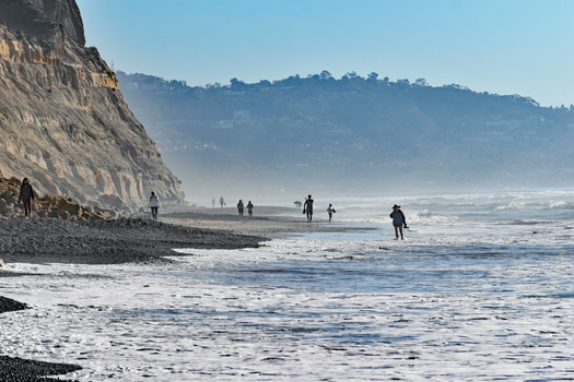 Surfers and beachgoers in the ocean with a tall cliff face rising behind them under clear skies.