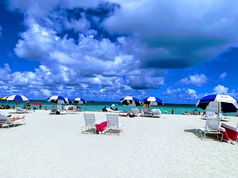 Sandy beach with lounge chairs and beach umbrellas under dramatic blue sky with white clouds.