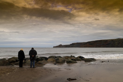 Two people standing in shallow water among rocks on a beach, facing the ocean under a dramatic cloudy sky.