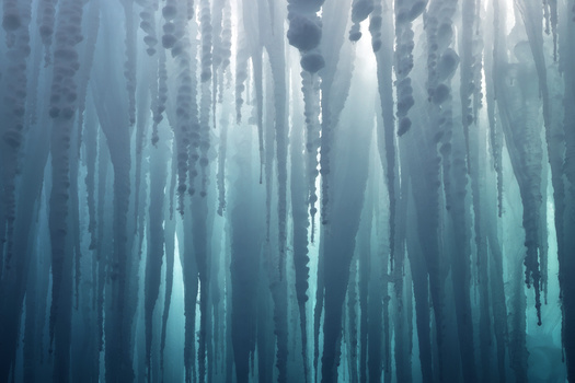 Vertical icicles hanging from a frozen overhang with misty, blue-toned backlighting creating an ethereal atmosphere.