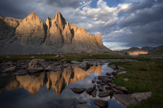 Dramatic rocky peaks reflected in still water during golden hour with storm clouds overhead.