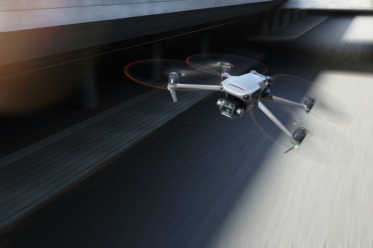 A compact quadcopter drone with folded arms sits on a dark wooden floor indoors, with a red cable visible in the background.