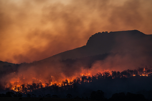 Wildfire burning across a forested mountainside silhouetted against an orange sky at dusk.