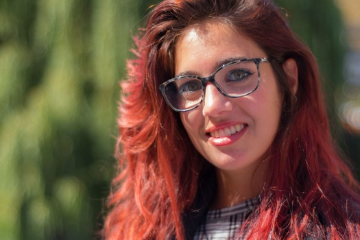 Woman with red hair and glasses smiling outdoors in natural sunlight.