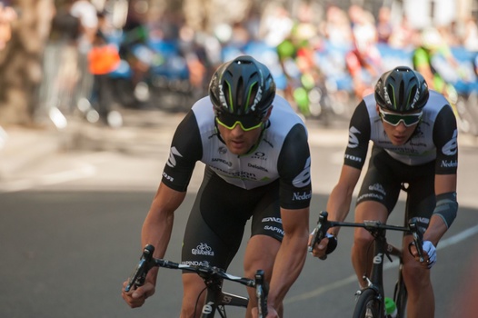 Two cyclists in black and white team kit riding side-by-side on a road with spectators blurred in the background.
