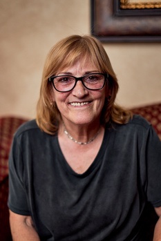 Woman wearing glasses and dark shirt smiling at camera in indoor setting.