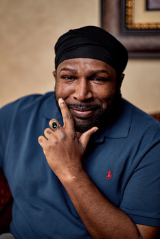 Man in blue polo shirt wearing black headband, smiling at camera with hand near face.