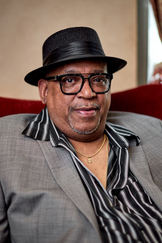 Portrait of a man wearing glasses, a black fedora hat, and striped shirt against a neutral backdrop.