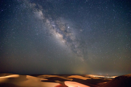 Milky Way galaxy arching across starry night sky above sand dunes illuminated by warm light.