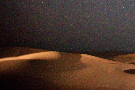 Desert sand dune silhouetted against a starry night sky with subtle warm lighting along the ridge.