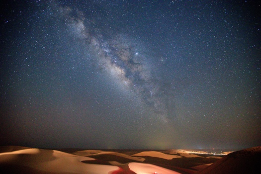 Milky Way galaxy arching across a night sky above desert sand dunes illuminated by warm light.