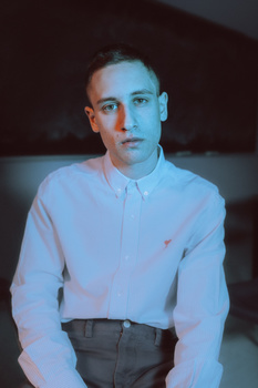 Man in light blue button-up shirt and dark trousers against dark background.