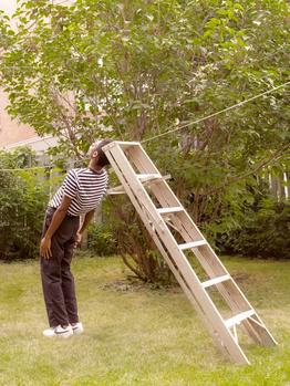 Person in striped shirt leaning against a wooden ladder in a garden setting with trees overhead.