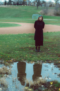 Woman in dark coat standing in field with puddle reflection in foreground.