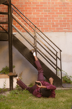 Person in burgundy clothing lying on grass beneath an industrial metal staircase against a brick wall.