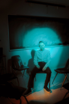 Man sitting in chair illuminated by cyan-blue projected light from a screen behind him.