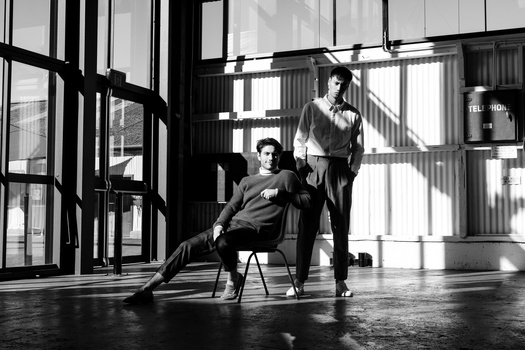 Two people posing in a modern industrial loft space with large windows and dramatic shadow patterns from the window frames.