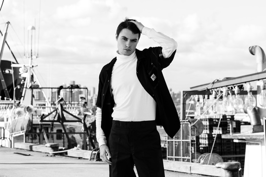 Man in black blazer and white shirt posing at a harbor with fishing boats in background.