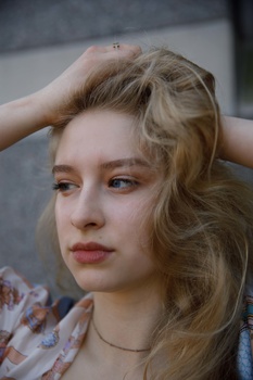 Young woman with blonde wavy hair posed against a gray background, hand raised to her head.