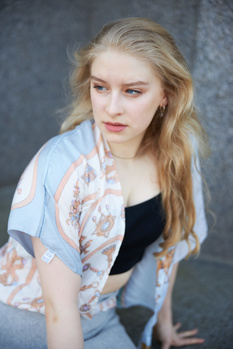 Young woman with long blonde hair wearing a patterned cardigan over a black top, posed against a textured gray backdrop.