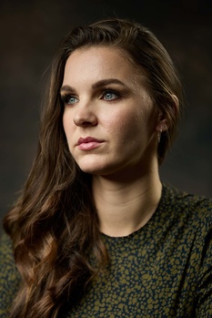 Woman with long brown hair wearing a textured green shirt, photographed against a dark background.