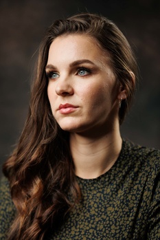 Woman with long dark hair wearing a textured dark shirt, photographed against a soft blurred background.