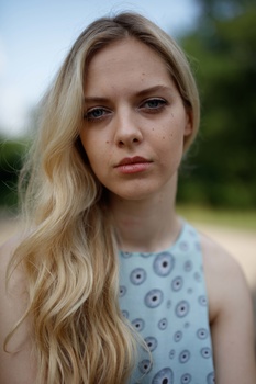Woman with long blonde hair wearing a light blue patterned sleeveless top, photographed outdoors with soft natural lighting.