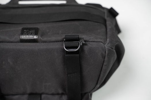 Close-up of black camera bag hardware showing metal D-ring attachment point and black webbing strap.